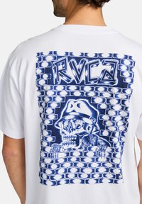 White t-shirt featuring a blue graphic print of a skull and graffiti-style text, set against a brick wall design.