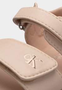Μπεζ λουράκια σανδαλιών με ραμμένα άκρα και μεταλλικό λογότυπο CK στο μπροστινό λουράκι, με ασημένιο κούμπωμα αγκράφας.