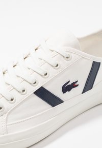 Valkoinen kangastus kenkä, jossa on merimiessinisiä nahkaosia, pyöreä kärki, litteät nauhat ja brodeerattu logo. Siinä on teksturoitu materiaali ja rei'ityksellä varustettua yksityiskohtaa.