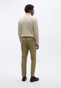 Ελαφρύ μπεζ ριμπ μπλουζάκι με ψηλό γιακά, συνδυασμένο με ανοιχτό ελιά παντελόνι, με τσέπες στο πίσω μέρος και μαύρα παπούτσια τύπου slip-on. Απλός, υφασμάτινος σχεδιασμός.