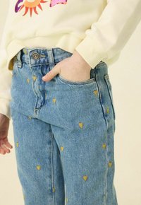 Jean bleu en denim avec de petits motifs de cœurs jaunes, dotés d'une poche avant et d'un bouton en métal. Partiellement recouvert par un sweat-shirt de couleur claire.
