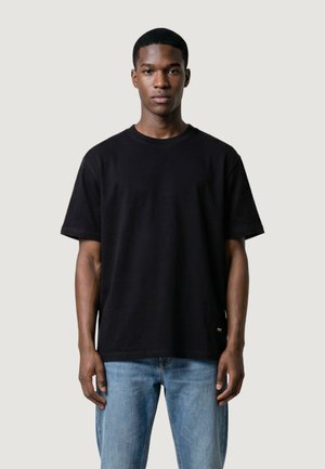 Giovane uomo in piedi, rivolto in avanti, indossa una maglietta nera a maniche corte semplice e jeans azzurri chiari su uno sfondo neutro.