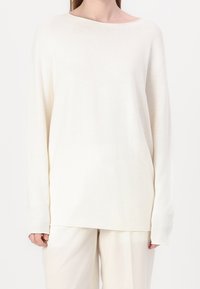 Biały sweter z długim rękawem o szerokim dekolcie, ściągaczach na mankietach i luźnym kroju. Materiał wydaje się miękki z subtelną fakturą. Połączony z jasnymi spodniami.