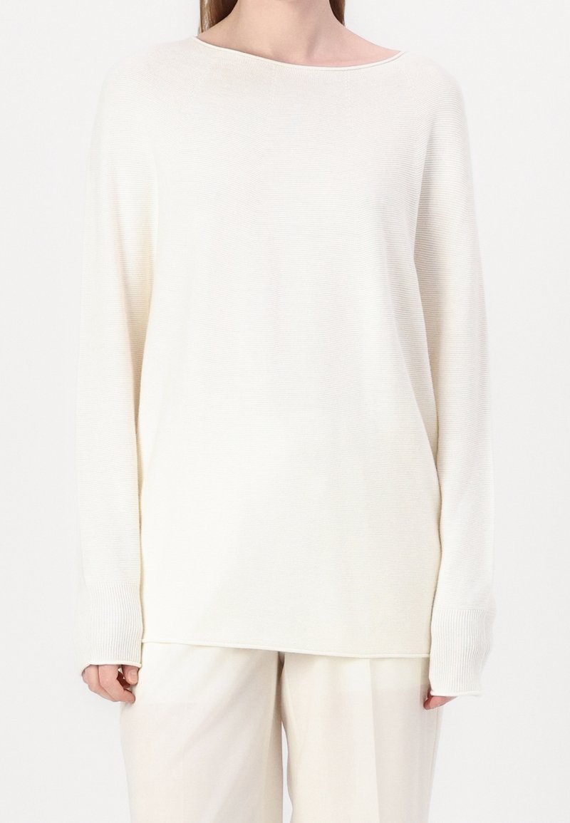 Biały sweter z długim rękawem o szerokim dekolcie, ściągaczach na mankietach i luźnym kroju. Materiał wydaje się miękki z subtelną fakturą. Połączony z jasnymi spodniami.