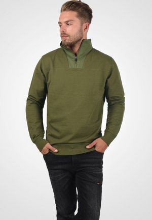 Olivengrønn genser med høy glidelås, lange ermer og rett snitt. Kombinert med slitte sorte jeans.