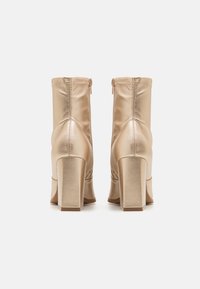 Bottes cheville en satin doré avec talon bloc, dotées d'une fermeture éclair, d'une texture lisse et d'un détail froncé à l'arrière.