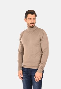 Maglione beige a collo alto in morbido materiale a maglia, caratterizzato da maniche lunghe e polsini a coste, abbinato a jeans scuri.