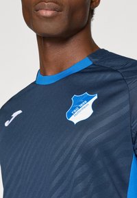 Maglia sportiva navy con colletto e dettagli blu. Presenta un tessuto testurizzato, il logo del club sul petto e una piccola grafica bianca.