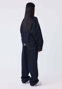 Conjunto de chaqueta de mezclilla azul marino de gran tamaño y pantalones con costuras visibles y etiqueta de logo en la parte de atrás. Completado con zapatillas deportivas blancas de suela gruesa.