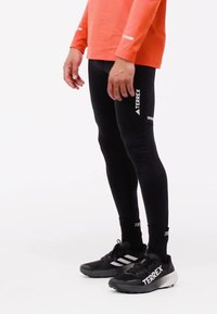 Person wearing black Terrex leggings, black Terrex sneakers with white soles, and an orange long-sleeve top standing on a white background.