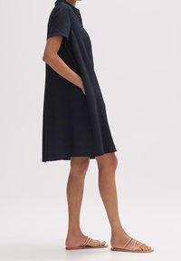 Marinblå kortärmad klänning med knappkrage, med en avslappnad passform, vertikal ribbad struktur och sidofickor. Matchas med platta sandaler.