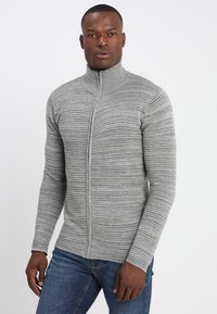 Grauer Reißverschluss-Pullover mit einem strukturierten, gerippten Design. Hoher Kragen und taillierte Form. Getragen mit blauen Jeans, was einen lässigen Look vermittelt.