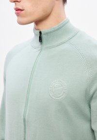 Φωτεινό πράσινο ζακέτα με φερμουάρ, κατασκευασμένη από υφασμάτινη υφή, με ψηλό γιακά, ριμπ λεπτομέρειες και κυκλικό κεντητό λογότυπο στο στήθος.