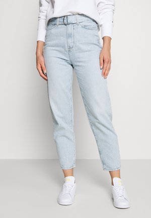 Person trägt hellblaue High-Waist-Mom-Jeans, weiße Sneaker und ein weißes langärmeliges Oberteil und steht vor einem schlichten hellen Hintergrund.