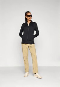 Mulher vestindo uma jaqueta preta com fecho, calças bege, ténis brancos e óculos escuros grandes e escuros, de pé com as mãos nos bolsos.