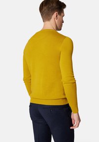 Musztardowy sweter z okrągłym dekoltem, z ribbowanymi mankietami i dołem. Gładka faktura, dopasowany krój, noszony na ciemnych spodniach.