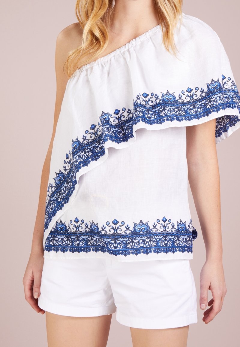 Vit off-the-shoulder topp med blå blommönster broderat längs fållen, med en volangkant och en lätt textur. Matchad med vita shorts.