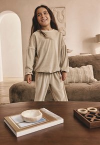 Completo beige a due pezzi con costine, con maniche larghe e pantaloni. Tessuto morbido, vestibilità comoda, indossato da un bambino in piedi all'interno vicino a un tavolo.