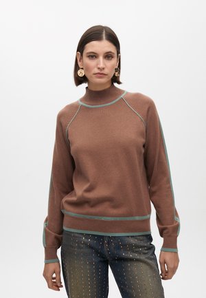 Pullover marrone con un colletto alto e accenti teal. Tessuto morbido, maniche raglan e polsini aderenti. Abbinato a jeans con dettagli metallici.