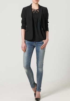 Blazer noir ajusté sur un top noir avec superposition de dentelle, associé à un jean skinny déchiré bleu clair et des chaussures à talons hauts noires.