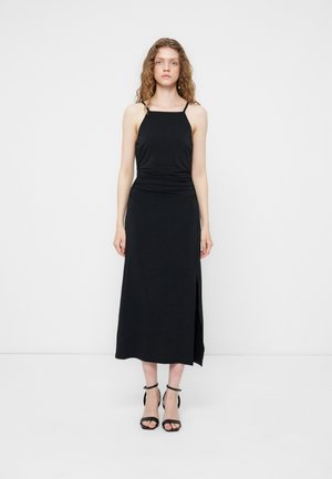 Γυναίκα με κατσαρά μαλλιά, φοράει μαύρο μίντι αμάνικο φόρεμα με άνοιγμα στο πλάι και μαύρα ψηλοτάκουνα σανδάλια, στέκεται μπροστά σε απλό λευκό φόντο.