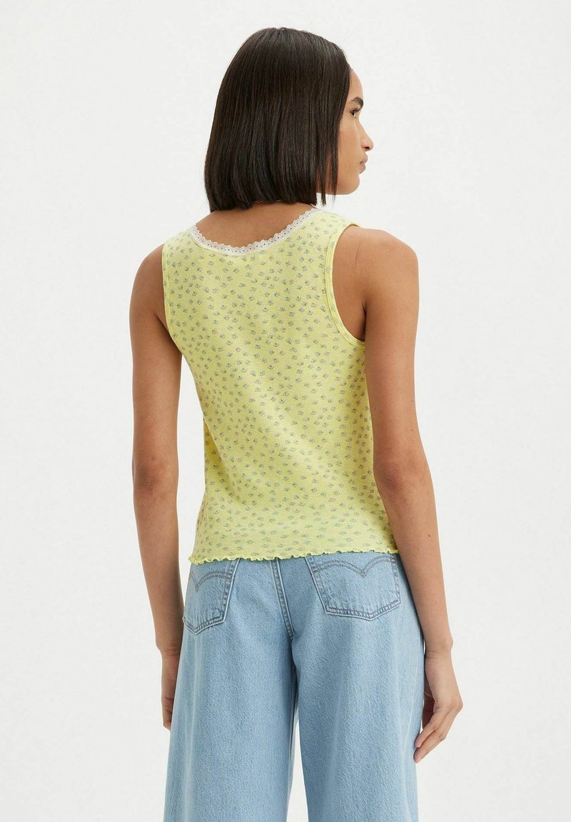 Top de tirantes amarillo con estampado floral y encaje en el escote, con un dobladillo en forma de onda, combinado con jeans de tiro alto en azul claro, visto desde la parte posterior.