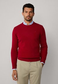 Homme portant un pull rouge à col rond sur une chemise boutonnée bleu clair avec un pantalon beige, se tenant d'une main dans sa poche contre un fond uni.