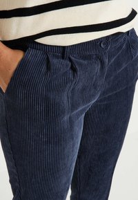 Pantalones de pana azul marino con rayas verticales. Incluyen características como bolsillos laterales, passadores para cinturón y cierre de botón. La textura es acanalada y suave.