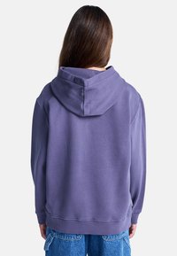 Lila Kapuzenpullover mit großer Kapuze, gerippten Bündchen und Saum. Weicher, strukturierter Baumwollstoff. Ein Etikett ist an der Seitennaht sichtbar.