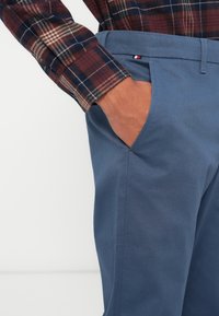 Pantaloni blu a trama con tasca laterale, abbinati a una camicia a maniche lunghe a quadri marrone e blu. Accento del marchio sottile sulla vita.