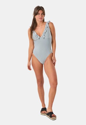 Kobieta stojąca, ubrana w prążkowany jednoczęściowy kostium kąpielowy z falbankami na ramiączkach oraz czarne sandały na platformie na jednolitym tle.