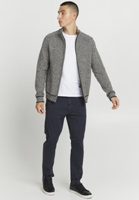 Giacca grigia con texture e zip alta, con zip marrone, indossata sopra una maglietta bianca, abbinata a pantaloni blu marino e sneaker nere.