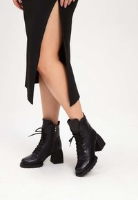 Jambes croisées portant une jupe noire fendue sur la cuisse et des bottines à talons noires à lacets sur un fond blanc.