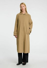 Tan lang frakke med krave, frontknapper og sidelommer; har løs pasform, lige ærmer og en glat stofstruktur.