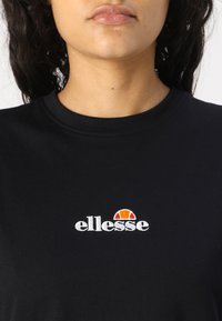 Juodas medvilninis marškinėliai su apykakle ir trumpomis rankovėmis, su balta "ellesse" logotipu ir virš jo oranžiniu pusapskretimu.