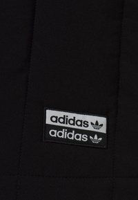 Černá tkanina má prošívaný design a zobrazuje dvě bílé nášivky loga Adidas s výraznými trojlístkovými akcenty.
