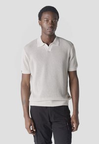 Jeune homme portant un polo en maille crème à manches courtes et un short cargo noir, debout devant un fond clair uni.