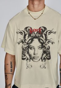 Crème-kleurige katoenen T-shirt met een grafische print van een vrouwelijk gezicht omringd door slangen, met daarboven rode tekst. Korte mouwen, ontspannen pasvorm.