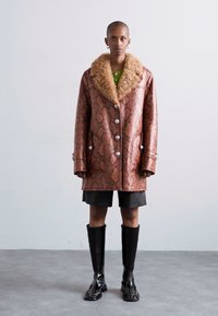Palton din piele sintetica maro cu un guler de blana bej, butoane mari si buzunare laterale, purtat cu pantaloni scurti negri si cizme glossy până la genunchi.