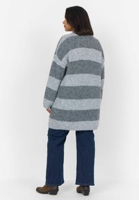 Pull en tricot oversize rayé gris et gris clair, avec un dos long et des poignets à côtes, associé à un jean bleu et des bottines marron.
