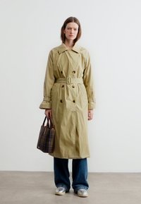 Femme portant un trench-coat beige ceinturé, un jean bleu, des chaussures argentées, et tenant un sac fourre-tout à carreaux marron, debout contre un mur blanc.