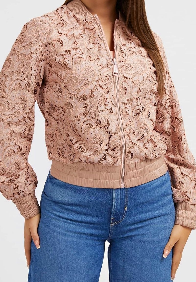 Veste bomber en dentelle rose à motifs floraux, fermeture éclair devant, poignets et taille côtelés, portée avec un jean bleu.