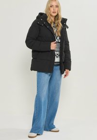 Giacca a vento nera con cappuccio, dotata di bottoni a pressione e texture trapuntata, indossata sopra un maglione a fantasia e abbinata a jeans a gamba larga.