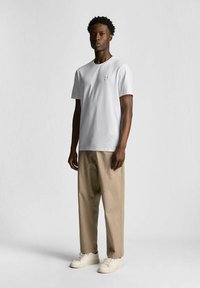T-shirt bianco in cotone con colletto rotondo e piccolo logo sul petto, abbinato a pantaloni beige e sneakers bianche.
