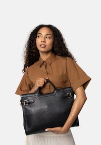 Sort læder tote bag med rektangulær form, to top-håndtag og guldfarvede spænde akcenter; holdes af en person iført en brun skjorte.