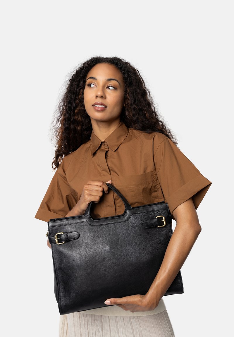 Sort læder tote bag med rektangulær form, to top-håndtag og guldfarvede spænde akcenter; holdes af en person iført en brun skjorte.