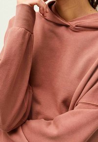 Sweat à capuche oversize en rose poudré, fabriqué en mélange de coton doux, avec poignets côtelés et coupe décontractée. Détail de couture visible.