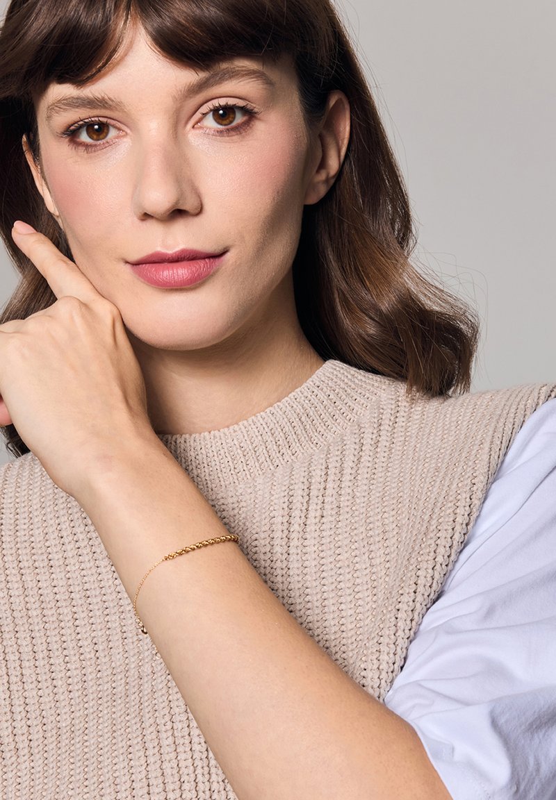 Złota bransoletka na nadgarstku, z delikatnym skręconym wzorem łańcuszka. Osoba nosi beżowy sweter z teksturowanym wzorem.