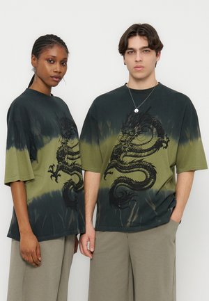 Doi adulți purtând cămăși supradimensionate asortate, în negru și verde măsliniu, cu imprimeu tie-dye și un grafic mare cu dragon negru, stând în fața unui fundal alb.