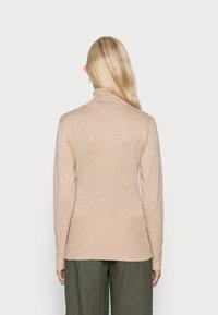 Femme aux longs cheveux blonds portant un pull à col roulé beige et un pantalon vert olive, vue de dos sur un fond uni.